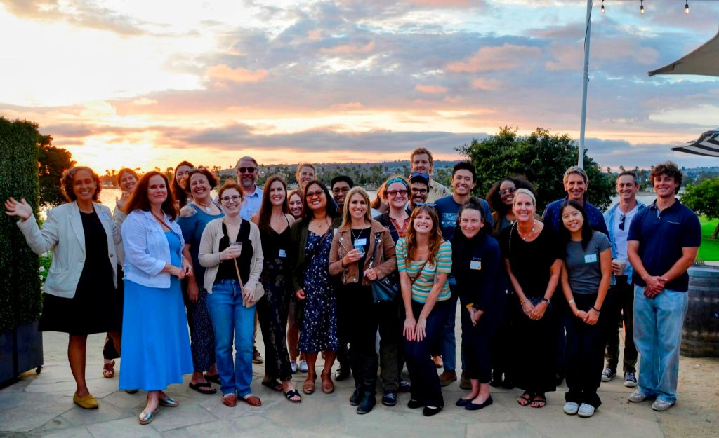 Group photo of around 20 San Diego members. They are posed outside, with a sunset in the background. Some are holding drinks. 