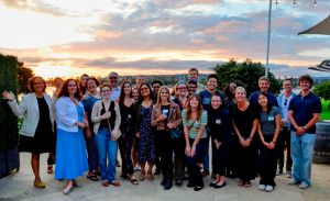 Group photo of around 20 San Diego members. They are posed outside, with a sunset in the background. Some are holding drinks.