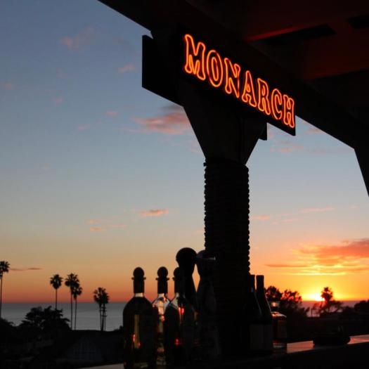 Photo of the Monarch sign. Sign is neon orange against a sunset background